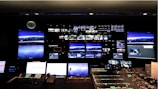 A control room with numerous screens displaying various images and streams on a large wall. Below the screens, a control panel with multiple buttons, switches, and telephones indicates a technical or broadcast setting. The room is dimly lit, and a clock is visible on the wall.