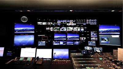 A control room with numerous screens displaying various images and streams on a large wall. Below the screens, a control panel with multiple buttons, switches, and telephones indicates a technical or broadcast setting. The room is dimly lit, and a clock is visible on the wall.