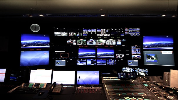 A control room with numerous screens displaying various images and streams on a large wall. Below the screens, a control panel with multiple buttons, switches, and telephones indicates a technical or broadcast setting. The room is dimly lit, and a clock is visible on the wall.