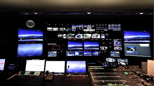 A control room with numerous screens displaying various images and streams on a large wall. Below the screens, a control panel with multiple buttons, switches, and telephones indicates a technical or broadcast setting. The room is dimly lit, and a clock is visible on the wall.