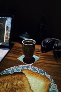 A dimly lit workspace featuring a laptop on the left with a cup of tea or coffee placed in the center on a coaster. Next to the cup, there are black headphones resting on the wooden desk. A patterned plate with two toasted sandwiches is partially visible at the bottom. In the background, a water bottle is slightly blurred.
