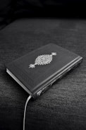 A closed book with an ornate cover design featuring decorative patterns and symbols, resting on a dark fabric surface. The book's cover is predominantly dark with intricate white artwork.