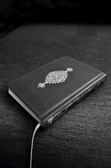 A closed book with an ornate cover design featuring decorative patterns and symbols, resting on a dark fabric surface. The book's cover is predominantly dark with intricate white artwork.