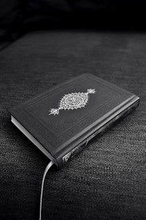 A closed book with an ornate cover design featuring decorative patterns and symbols, resting on a dark fabric surface. The book's cover is predominantly dark with intricate white artwork.