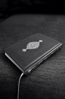 A closed book with an ornate cover design featuring decorative patterns and symbols, resting on a dark fabric surface. The book's cover is predominantly dark with intricate white artwork.
