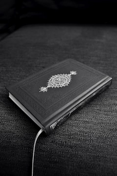 A closed book with an ornate cover design featuring decorative patterns and symbols, resting on a dark fabric surface. The book's cover is predominantly dark with intricate white artwork.