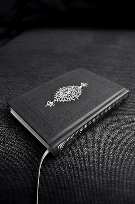 A closed book with an ornate cover design featuring decorative patterns and symbols, resting on a dark fabric surface. The book's cover is predominantly dark with intricate white artwork.