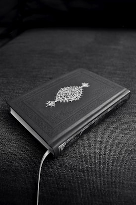 A closed book with an ornate cover design featuring decorative patterns and symbols, resting on a dark fabric surface. The book's cover is predominantly dark with intricate white artwork.