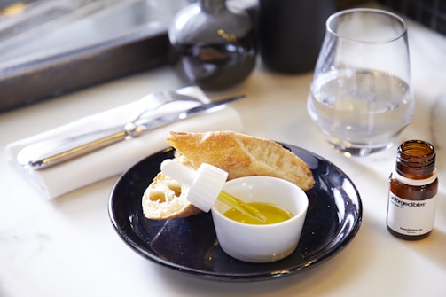 A table setting with a black plate holding sliced bread, a small bowl of olive oil with a dropper, and a glass bottle labeled 'Introgredibles.' A napkin with a fork and butter knife is set beside a clear glass of water.