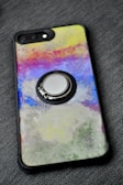 A smartphone case with a colorful abstract design featuring a mix of vibrant colors including yellow, pink, blue, and green. The design has a textured, painted look. The case also has a metallic ring holder attached to its back.