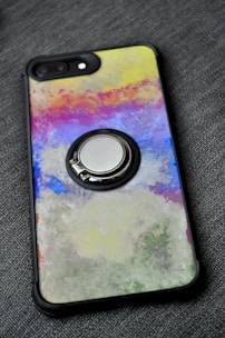 A smartphone case with a colorful abstract design featuring a mix of vibrant colors including yellow, pink, blue, and green. The design has a textured, painted look. The case also has a metallic ring holder attached to its back.