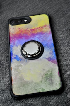 A smartphone case with a colorful abstract design featuring a mix of vibrant colors including yellow, pink, blue, and green. The design has a textured, painted look. The case also has a metallic ring holder attached to its back.