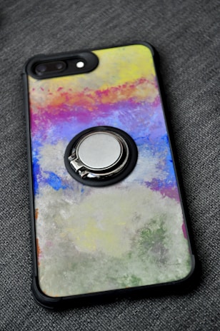 A smartphone case with a colorful abstract design featuring a mix of vibrant colors including yellow, pink, blue, and green. The design has a textured, painted look. The case also has a metallic ring holder attached to its back.