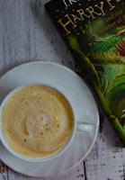 A cup of frothy cappuccino is placed on a white saucer on a rustic wooden surface. Next to the cup is a partially visible book with a green and gold cover, featuring an illustration of a serpent.