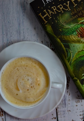 A cup of frothy cappuccino is placed on a white saucer on a rustic wooden surface. Next to the cup is a partially visible book with a green and gold cover, featuring an illustration of a serpent.