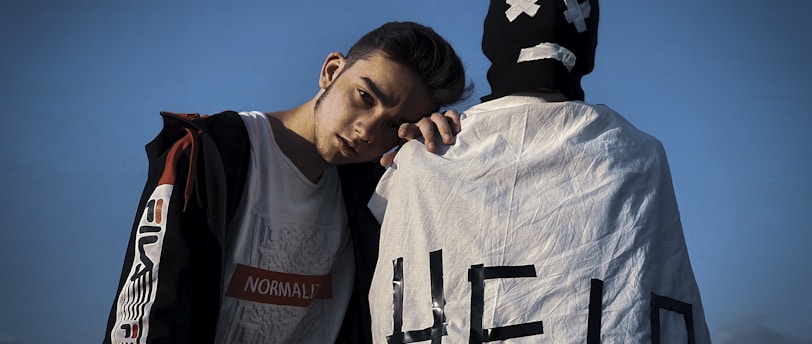 A young person stands leaning on someone dressed in a white cloth with 'HELP' written on it, wearing a mask with a sad face. The sky is clear and blue in the background.