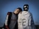 A young person stands leaning on someone dressed in a white cloth with 'HELP' written on it, wearing a mask with a sad face. The sky is clear and blue in the background.