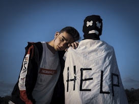 A young person stands leaning on someone dressed in a white cloth with 'HELP' written on it, wearing a mask with a sad face. The sky is clear and blue in the background.
