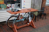A creatively repurposed bicycle is used as a unique outdoor table setup, placed on a brick sidewalk. The bicycle has a wooden tabletop mounted on it, and on the tabletop are various objects including two large metal cans and a roll of white paper. Behind the table is a window decorated with colorful holiday lights and signs advertising hot dogs and urging customers to eat there. Two black metal chairs and a small table are positioned nearby.