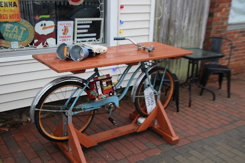 A creatively repurposed bicycle is used as a unique outdoor table setup, placed on a brick sidewalk. The bicycle has a wooden tabletop mounted on it, and on the tabletop are various objects including two large metal cans and a roll of white paper. Behind the table is a window decorated with colorful holiday lights and signs advertising hot dogs and urging customers to eat there. Two black metal chairs and a small table are positioned nearby.