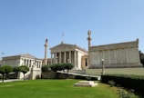 A classical Greek-style building with columns and ornate statues, surrounded by manicured green lawns and hedges. The sky is clear and blue, highlighting the architectural details of the structure.
