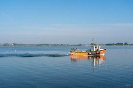 A small fishing boat with orange and black colors is moving across a calm lake. The boat is making gentle waves in the otherwise still water. In the distance, there is a shoreline with trees and buildings visible under a clear blue sky.