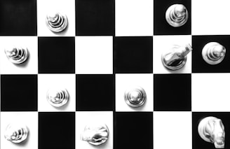 A black and white chessboard with a few silver chess pieces placed strategically across the squares. The chess pieces appear to be metallic, reflecting light and creating a shiny appearance. The contrast between the black and white squares enhances the visual appeal.