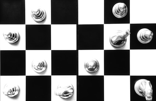 A black and white chessboard with a few silver chess pieces placed strategically across the squares. The chess pieces appear to be metallic, reflecting light and creating a shiny appearance. The contrast between the black and white squares enhances the visual appeal.