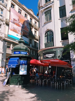 A bustling urban scene with people sitting under red umbrellas at an outdoor seating area near a kebab shop. A large advertisement is displayed on a tall building to the left, and there is a tourism information kiosk in the foreground. The buildings have a classic architecture with large windows and detailed facades.
