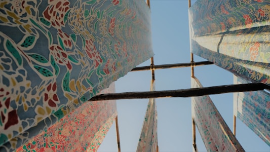 Colorful patterned fabrics are hung on wooden poles, drying in the open air under a clear blue sky. The fabrics feature intricate floral designs with various colors creating a vibrant contrast.
