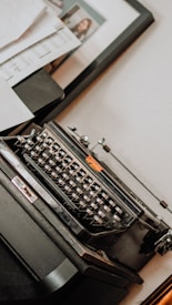 A vintage typewriter positioned on a wooden surface, surrounded by scattered papers and a framed photograph in the background. The typewriter has a classic black design with metallic keys and an orange logo. Soft lighting creates a nostalgic atmosphere.