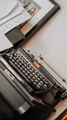 A vintage typewriter positioned on a wooden surface, surrounded by scattered papers and a framed photograph in the background. The typewriter has a classic black design with metallic keys and an orange logo. Soft lighting creates a nostalgic atmosphere.