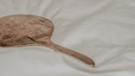 A wooden paddle with a worn and textured surface is resting on a soft, cream-colored fabric. The paddle features an engraved circular emblem near its center. The fabric has subtle folds, creating a gentle background.