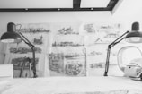 Detailed architectural sketches and blueprints cover a wall, illuminated by two large black desk lamps on either side of a white workspace. The sketches depict different architectural structures and landscapes, showcasing intricate detail and artistic skill. The scene has a monochromatic color scheme, emphasizing the focus on the drawings and the creative process.