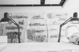 Detailed architectural sketches and blueprints cover a wall, illuminated by two large black desk lamps on either side of a white workspace. The sketches depict different architectural structures and landscapes, showcasing intricate detail and artistic skill. The scene has a monochromatic color scheme, emphasizing the focus on the drawings and the creative process.