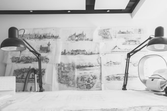 Detailed architectural sketches and blueprints cover a wall, illuminated by two large black desk lamps on either side of a white workspace. The sketches depict different architectural structures and landscapes, showcasing intricate detail and artistic skill. The scene has a monochromatic color scheme, emphasizing the focus on the drawings and the creative process.