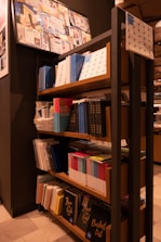 A bookshelf filled with various notebooks and books, organized neatly. The shelves contain items of different sizes and colors, with prominent blue and colorful book spines. Above the shelf, a bulletin board displays a collage of images and papers. The setting appears to be a stationary or bookshop, with warm lighting and a modern interior design.