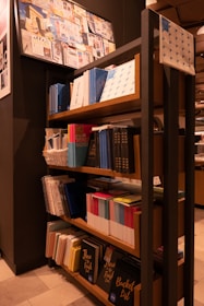 A bookshelf filled with various notebooks and books, organized neatly. The shelves contain items of different sizes and colors, with prominent blue and colorful book spines. Above the shelf, a bulletin board displays a collage of images and papers. The setting appears to be a stationary or bookshop, with warm lighting and a modern interior design.