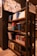 A bookshelf filled with various notebooks and books, organized neatly. The shelves contain items of different sizes and colors, with prominent blue and colorful book spines. Above the shelf, a bulletin board displays a collage of images and papers. The setting appears to be a stationary or bookshop, with warm lighting and a modern interior design.