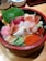 A colorful bowl of sashimi and other ingredients arranged over rice. Pieces of raw fish, shrimp, fish roe, sliced cucumber, and a green leaf are visible, creating a vibrant and appetizing appearance.