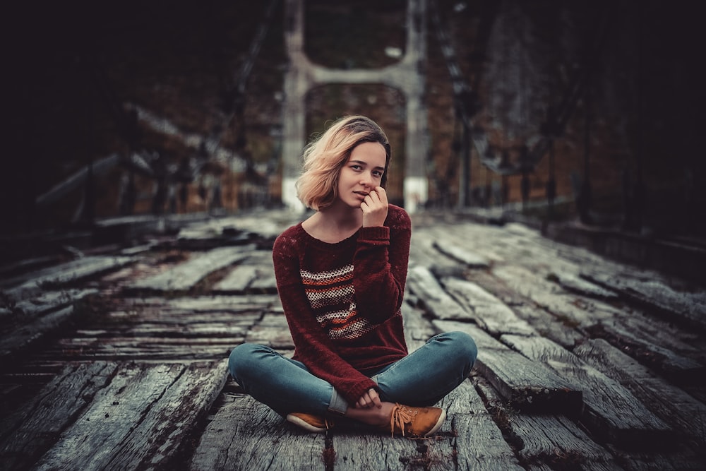 woman in red long sleeve shirt and blue denim jeans sitting on brown wooden pathway during