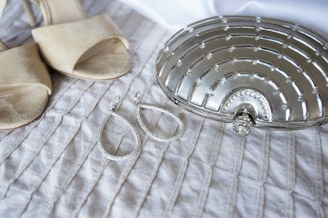 A pair of beige high-heeled sandals and two large teardrop-shaped earrings are placed on a textured white fabric. Beside them lies a metallic silver clutch with a decorative, geometric pattern.