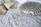 A pair of beige high-heeled sandals and two large teardrop-shaped earrings are placed on a textured white fabric. Beside them lies a metallic silver clutch with a decorative, geometric pattern.
