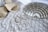 A pair of beige high-heeled sandals and two large teardrop-shaped earrings are placed on a textured white fabric. Beside them lies a metallic silver clutch with a decorative, geometric pattern.