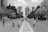 Children are riding bicycles down a wide, empty city street lined with trees and tall buildings. The scene captures a lively family-friendly atmosphere with pedestrians walking alongside. The monochrome coloring gives a timeless and classic feel.