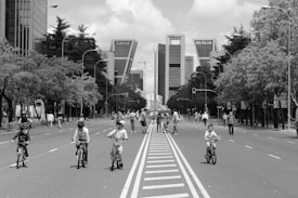 Children are riding bicycles down a wide, empty city street lined with trees and tall buildings. The scene captures a lively family-friendly atmosphere with pedestrians walking alongside. The monochrome coloring gives a timeless and classic feel.