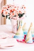 A vibrant and festive table arrangement features colorful party hats, flowers in a shiny silver pot, and neatly stacked plates and napkins. The soft pink flowers contrast with the multi-colored party hats, creating a lively, celebratory atmosphere.