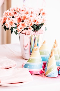 A vibrant and festive table arrangement features colorful party hats, flowers in a shiny silver pot, and neatly stacked plates and napkins. The soft pink flowers contrast with the multi-colored party hats, creating a lively, celebratory atmosphere.