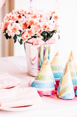 A vibrant and festive table arrangement features colorful party hats, flowers in a shiny silver pot, and neatly stacked plates and napkins. The soft pink flowers contrast with the multi-colored party hats, creating a lively, celebratory atmosphere.
