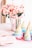 A vibrant and festive table arrangement features colorful party hats, flowers in a shiny silver pot, and neatly stacked plates and napkins. The soft pink flowers contrast with the multi-colored party hats, creating a lively, celebratory atmosphere.